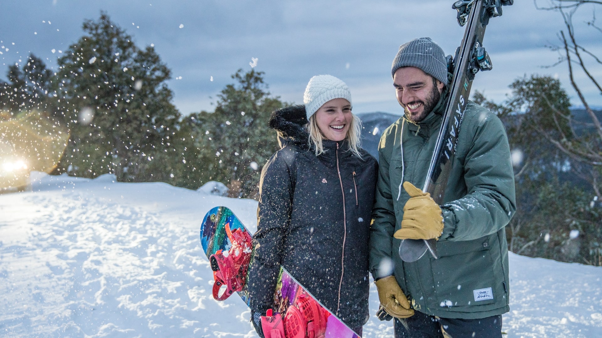Man and woman in snow gear holding a snowboard and skiis